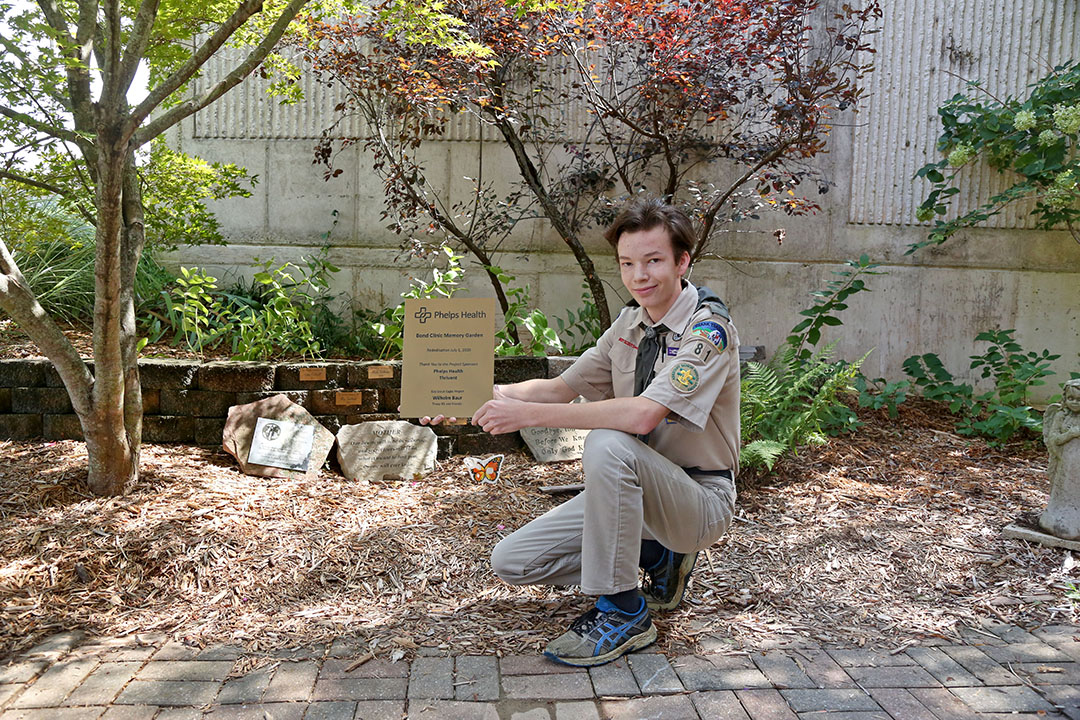 Scout WIl Baur holding a plaque in front of Bond Memorial Garden