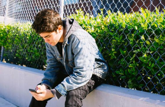 A teen boy sits next to a fence and bush looking at his phone